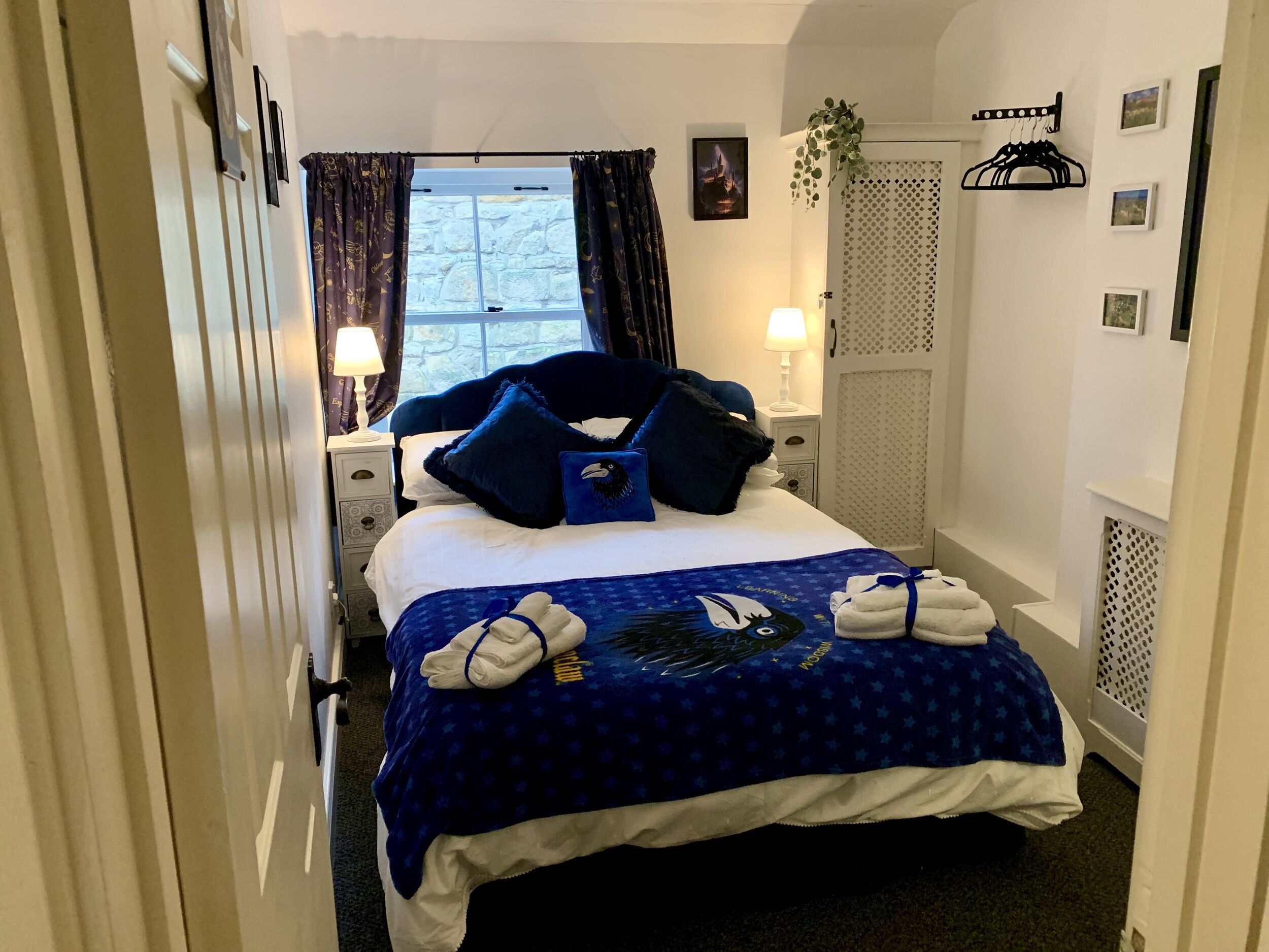 A hotel room with a bed, a window with curtains, a nightstand with a lamp, and a wall-mounted coat rack.