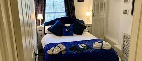 A hotel room with a bed, a window with curtains, a nightstand with a lamp, and a wall-mounted coat rack.