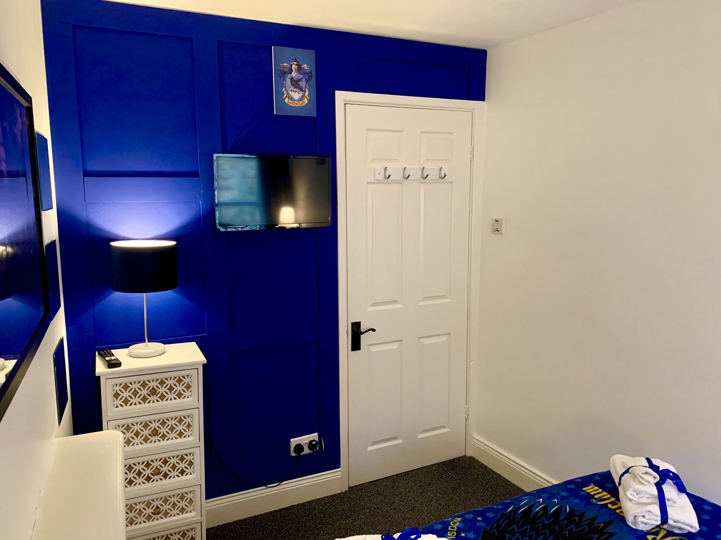 Une pièce avec un mur d’accent bleu, une porte blanche avec un porte-manteau, un t éléviseur à écran plat fixé au-dessus de la porte, une table de chevet blanche avec une lampe, et un lit recouvert d’une couvre-lit à motifs bleus et jaunes.