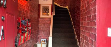 A narrow hallway with red brick walls, a red door, and a staircase leading to an upper floor.