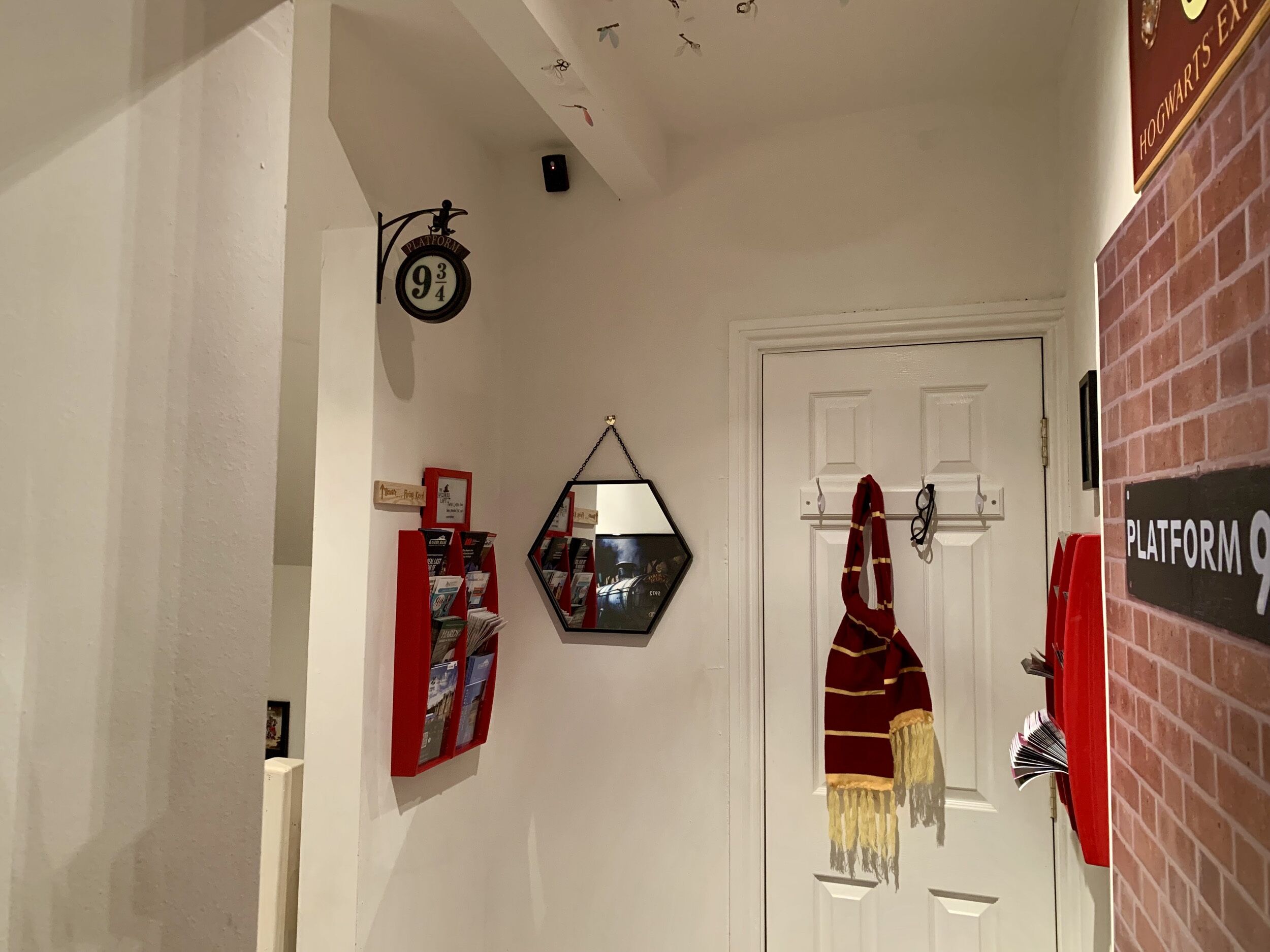 A hallway with a white door, a red and gold striped scarf hanging on the door, a red bookcase, and a sign reading "Platform 9 3/4".