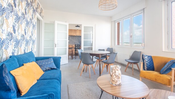 A modern living room with a blue sofa, yellow cushions, a wooden coffee table, and a yellow armchair. There is a dining area with a round table and chairs. The room has large windows and a view of the outside. - Banc des Flandres - Appt proche plage (Bray-Dunes)