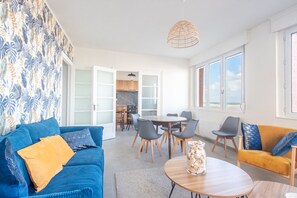 A modern living room with a blue sofa, yellow cushions, a wooden coffee table, and a yellow armchair. There is a dining area with a round table and chairs. The room has large windows and a view of the outside.