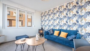 A modern living room with a blue sofa, two round wooden tables, a decorative vase, and a patterned wallpaper. - Banc des Flandres - Appt proche plage (Bray-Dunes)