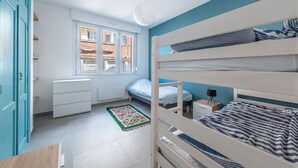 A bedroom with a bunk bed, a window with blinds, a radiator, a desk with a lamp, and a patterned rug. - Banc des Flandres - Appt proche plage (Bray-Dunes)