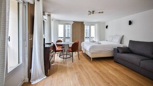 A modern bedroom with a bed, a gray sofa, a dining table with chairs, and a view of the outside through large windows. - 33.Studio 4Pers#Madeleine#Gallerie La Fayette (Paris)