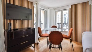 A modern hotel room with a flat-screen TV, a dark wooden dresser, a glass dining table with orange chairs, and large windows offering a view of the city. - 33.Studio 4Pers#Madeleine#Gallerie La Fayette (Paris)