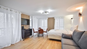 A modern hotel room with a bed, a sofa, a TV, a desk with chairs, and a wooden floor. - 33.Studio 4Pers#Madeleine#Gallerie La Fayette (Paris)