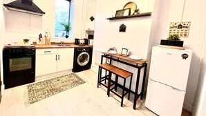 Eine kleine Küche mit weißen Schränken, einem schwarzen Herd, einer Waschmaschine, einer hölzernen Sitzbank und einem weißen Kühlschrank.