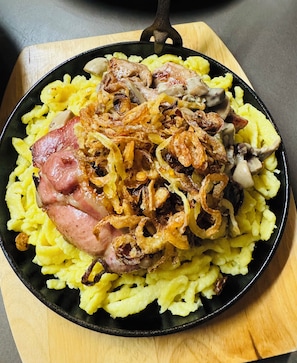 A skillet with food, including ham and mushrooms, on a wooden board. - Hotel-Restaurant Maien (Todtmoos)