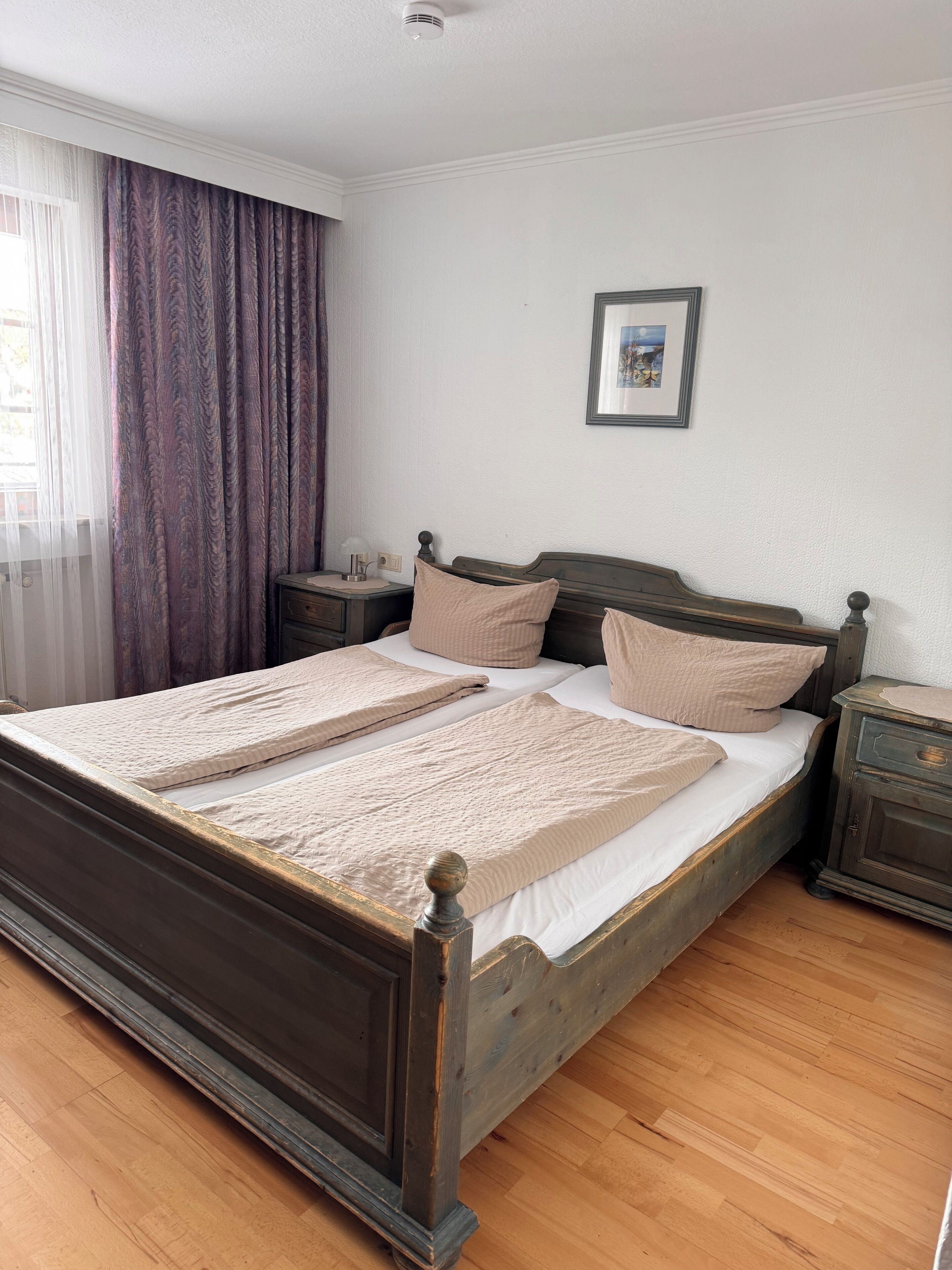 Un dormitorio con una cama de madera, una mesita de noche, un cuadro en la pared y una ventana con cortinas.