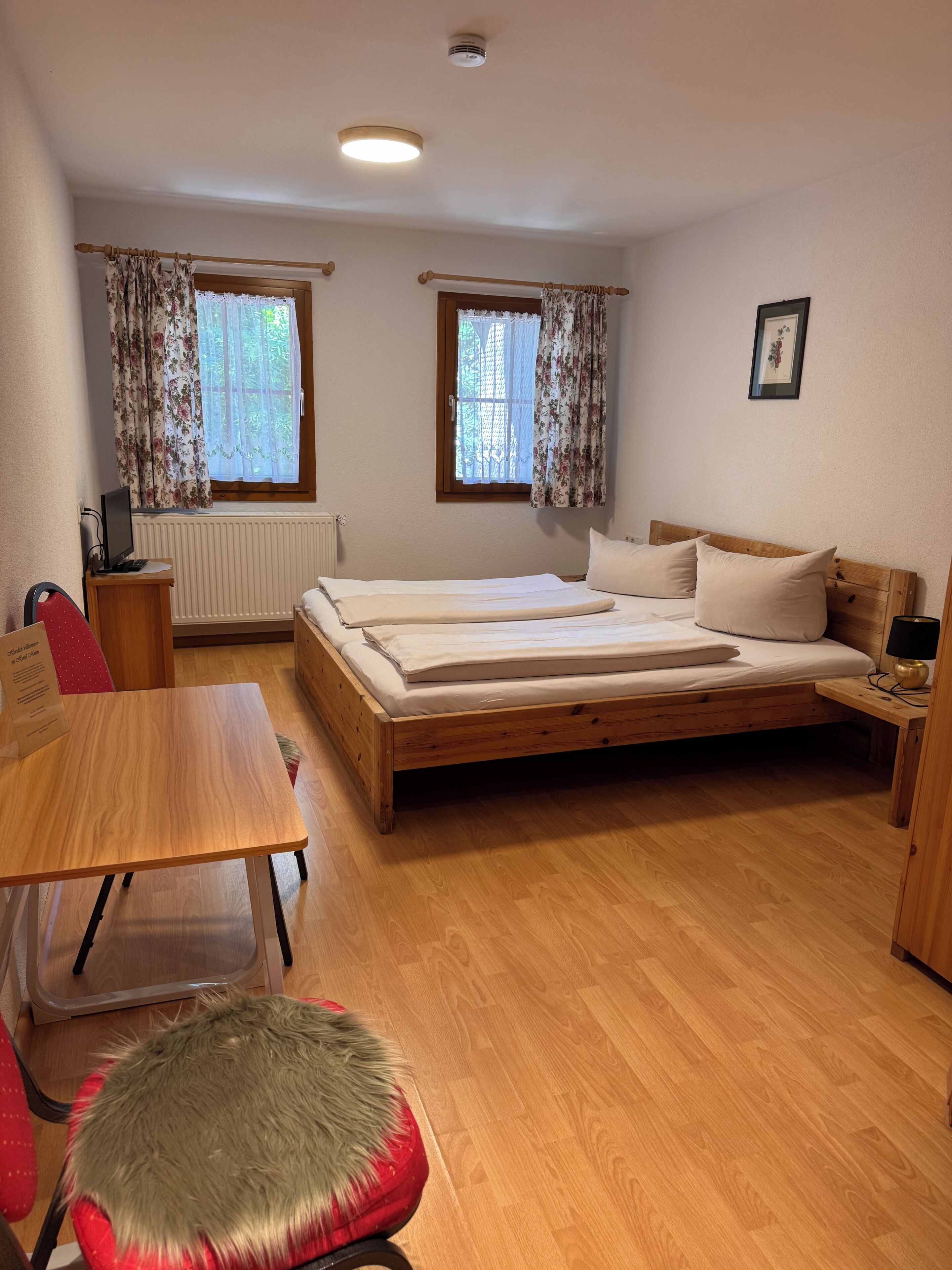 Ein Schlafzimmer mit einem hölzernen Bett, zwei mit Vorhängen versehenen Fenstern, einem Schreibtisch, einem Stuhl und einer Heizung.