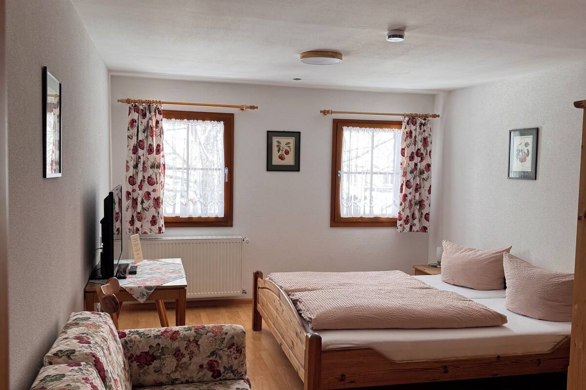 A bedroom with a wooden bed, a floral sofa, two windows with curtains, a bedside table, and a radiator.