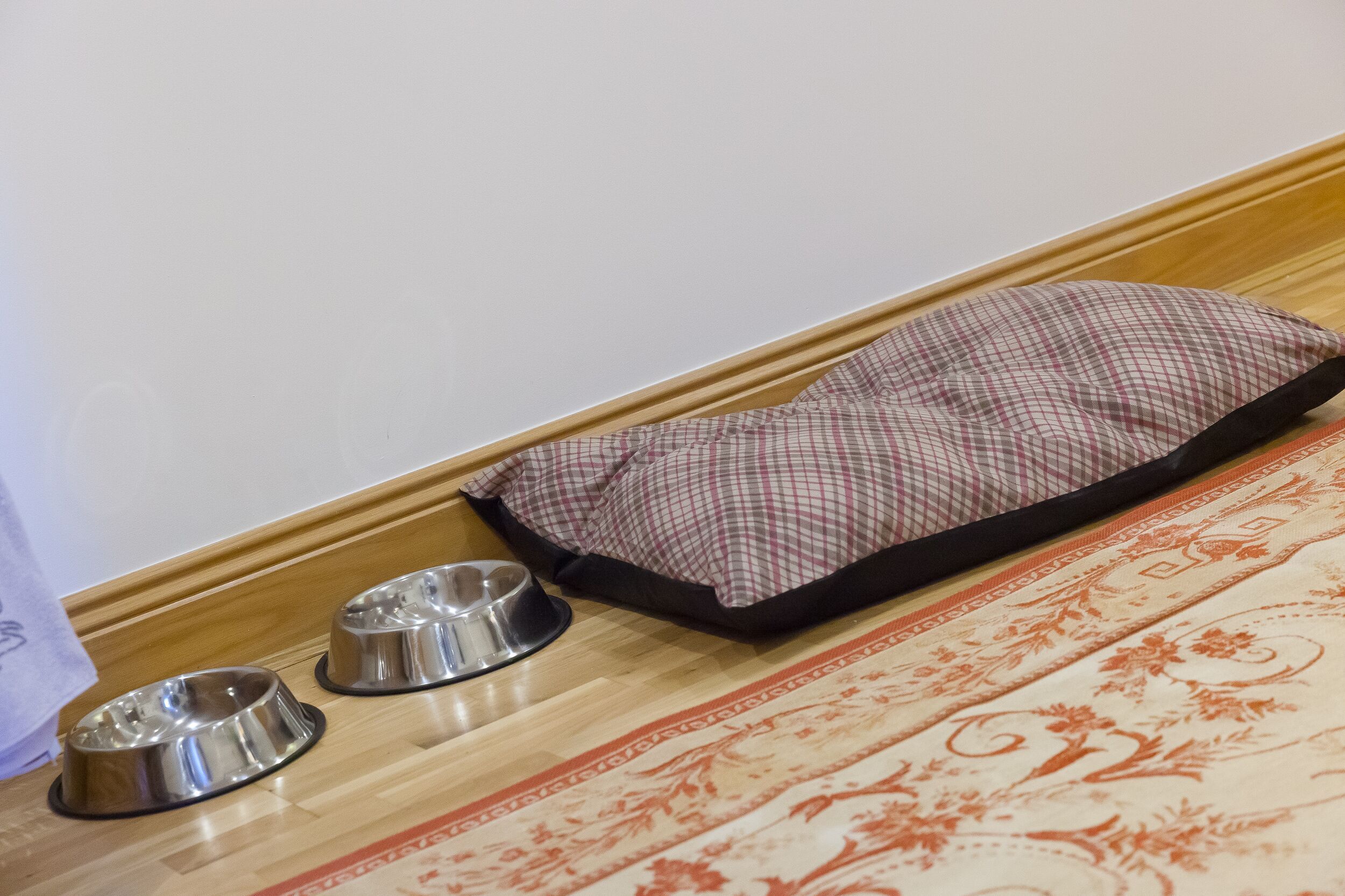 A pet bed and two metal bowls on a wooden floor with a patterned rug.