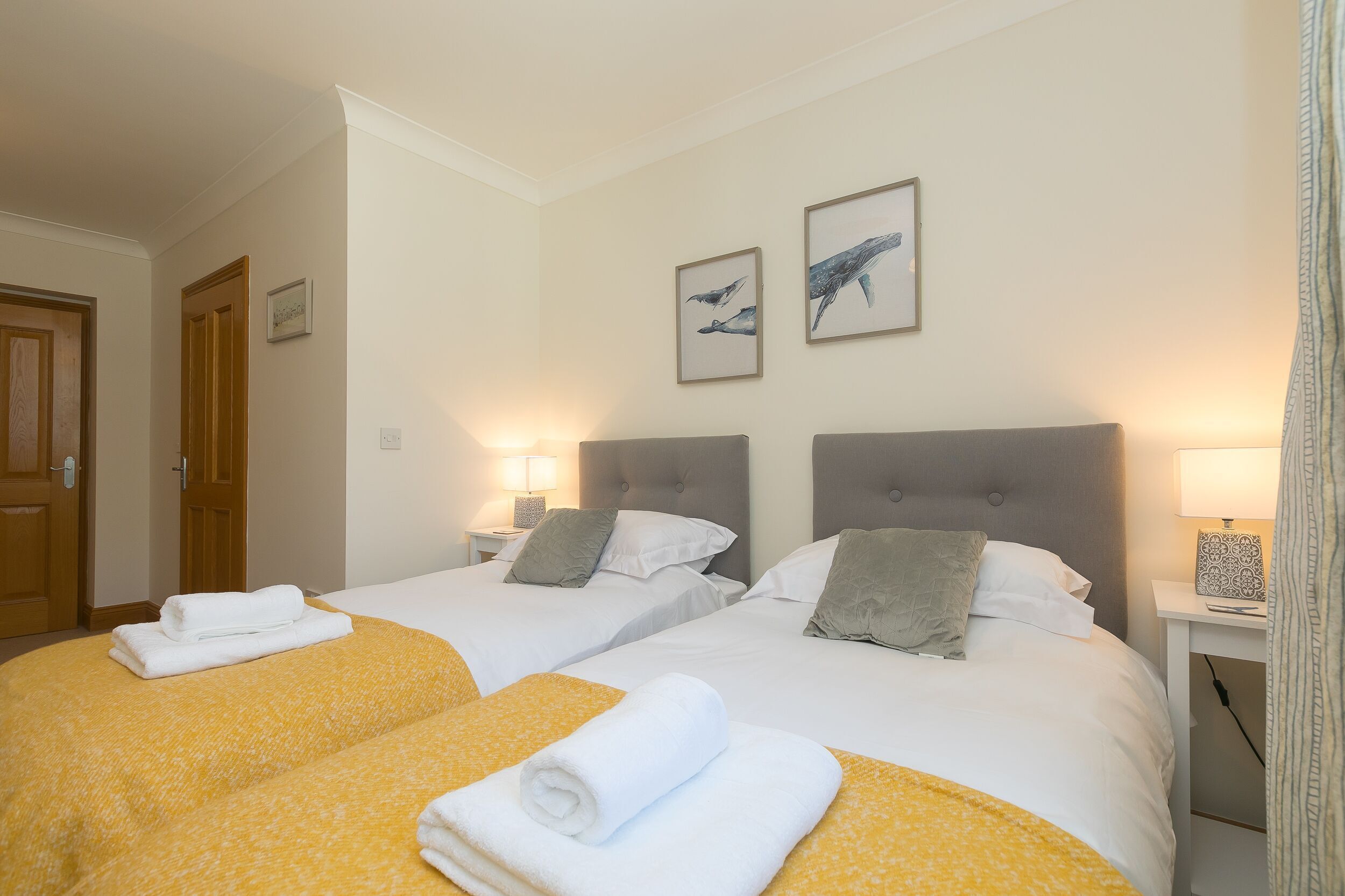 A hotel room with two beds, each with a headboard, white linens, and a yellow patterned bedspread. There are two wall-mounted lamps on either side of the beds, and a framed picture of whales above them.