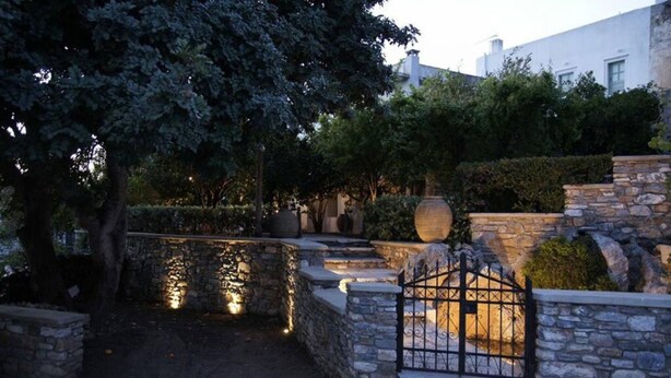 A stone wall with a metal gate, a pathway leading to a building, and a tree. - Giannakis Filoxenia (Naxos)