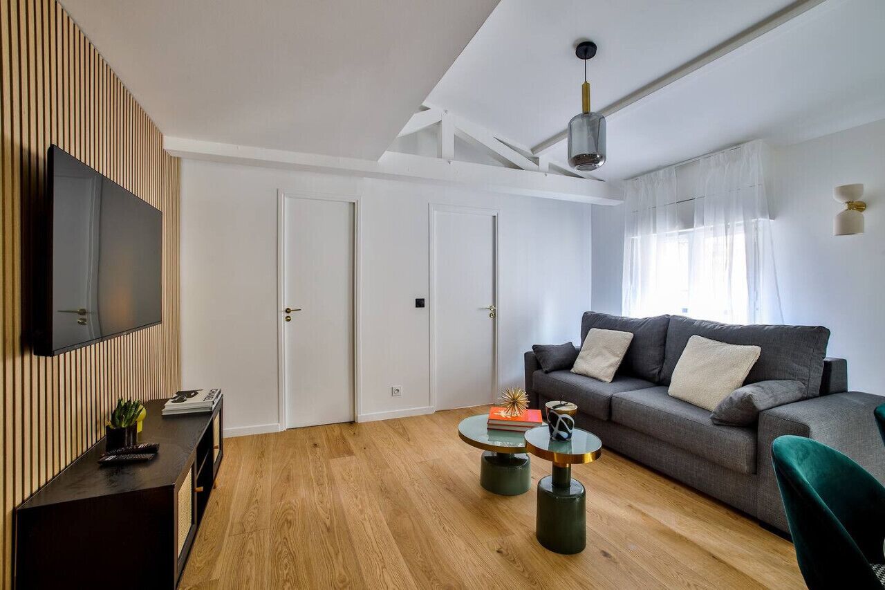 A modern living room with a grey sofa, a coffee table, a TV on a wooden panel wall, and a ceiling light.