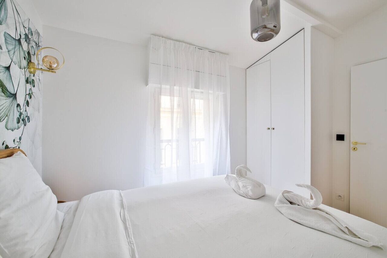 A hotel room with a bed, a wardrobe, a window with sheer curtains, and a wall-mounted light fixture.