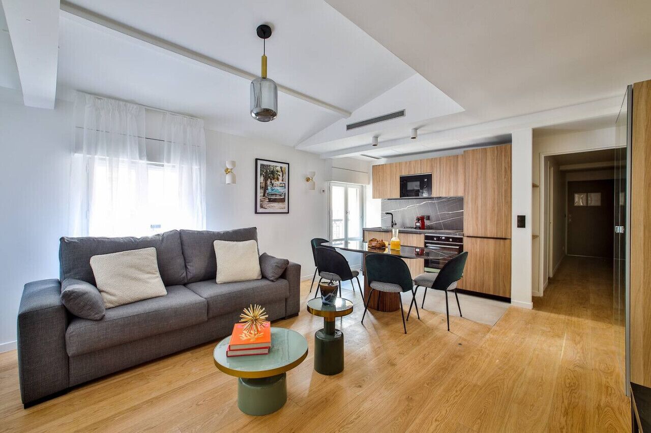 A modern living room with a grey sofa, a small round table with a book, and a dining area with chairs. A kitchen with wooden cabinets and a microwave is visible in the background.