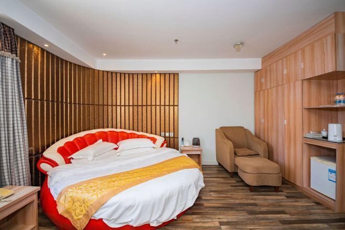 A modern hotel room with a large bed, a wooden panel wall, a wooden wardrobe, and a small desk with a lamp.