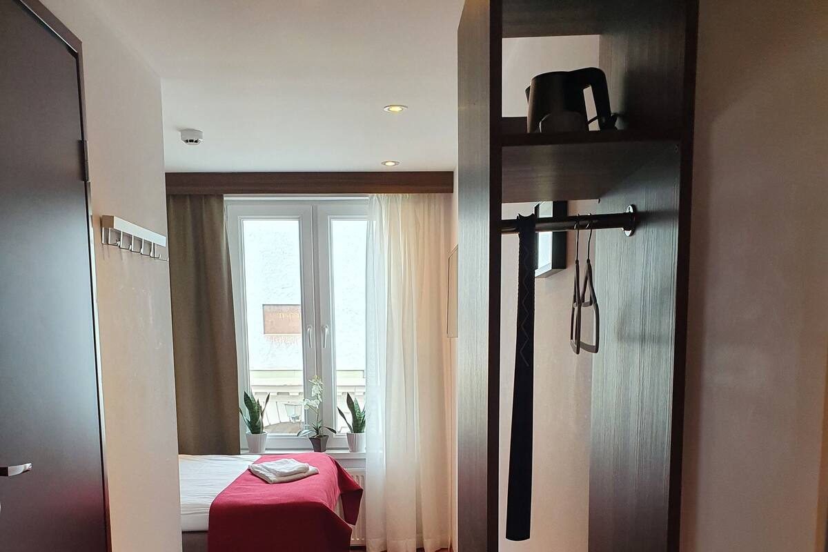 A hotel room with a bed, a small table, a window with curtains, a wooden floor, and a coat rack.