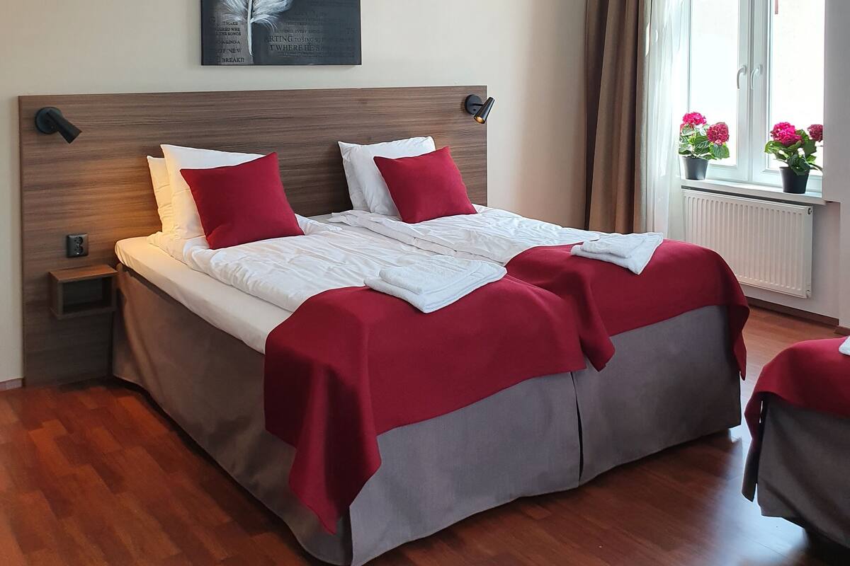 A neatly made bed with red and white bedding, a feather artwork on the wall, and a window with curtains.