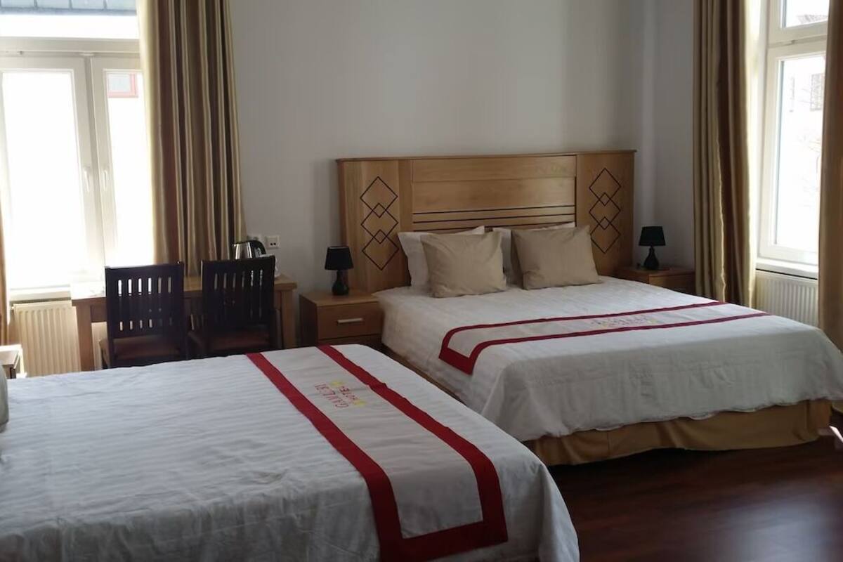 A hotel room with two beds, a wooden headboard, bedside tables with lamps, a chair, and a window with curtains.