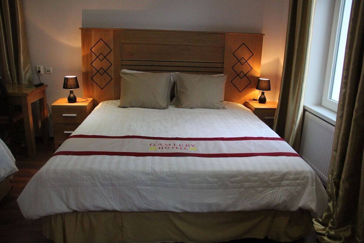 A hotel room with a bed, two bedside tables with lamps, a wooden headboard, and a window with curtains.