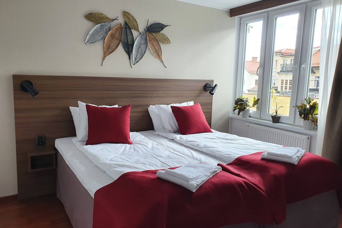 A neatly made bed with white and red bedding, a wooden headboard, and decorative leaf wall art.