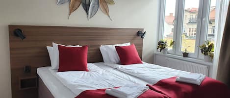 A neatly made bed with white and red bedding, a wooden headboard, and decorative leaf wall art.
