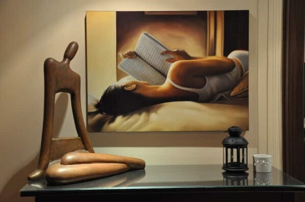Un tableau représentant une personne lisant sur un lit, une sculpture en bois et une petite table sur laquelle se trouvent une lanterne et un verre.