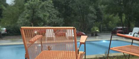 Una silla de mimbre junto a la piscina, con vistas a árboles y un coche estacionado al fondo.