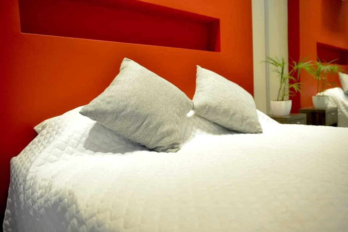 Una cama con ropa de cama blanca y dos almohadas, una pared roja y una planta en maceta sobre una mesita de noche.