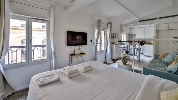 A hotel room with a large bed, a TV, a balcony with a railing, and a kitchenette area.