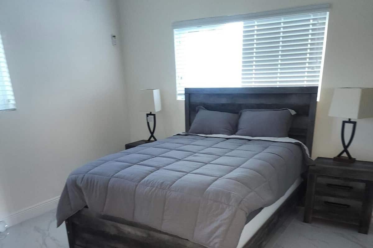 Un dormitorio con una cama, mesitas de noche, una ventana con persianas y un equipo de aire acondicionado en la pared.