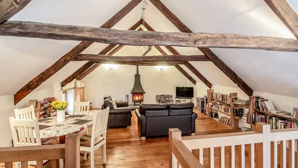 A cozy attic room with exposed wooden beams, a fireplace, a sofa, a dining table with chairs, and a bookshelf.