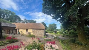 A rustic stone building with a thatched roof, surrounded by a well-maintained garden with flowering plants.