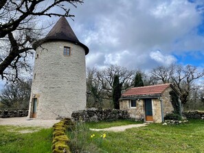 Ein steinerner Turm mit einer Kegeldach, ein kleines Steinhaus mit rotem Ziegeldach und eine Wiese mit gelben Blumen.
