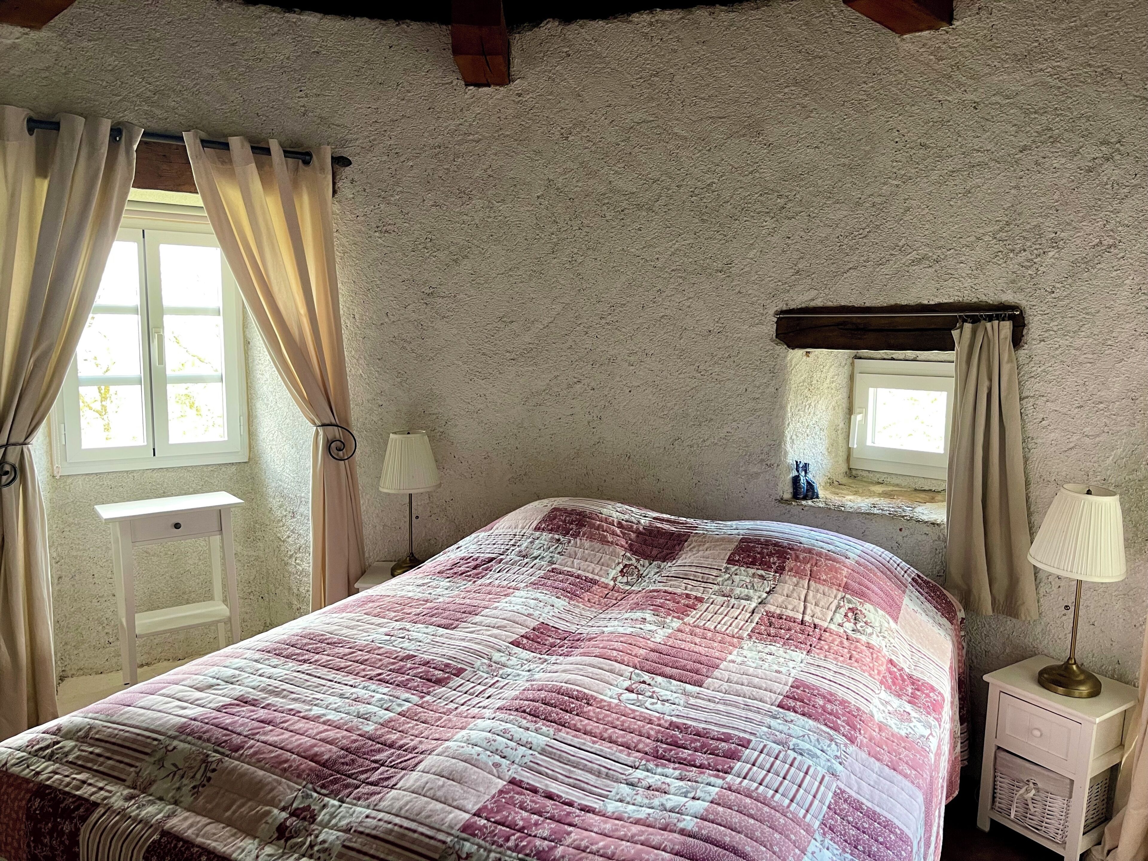 Ein Schlafzimmer mit einem Bett, einem Nachttisch, einer Lampe, einem Fenster mit Vorhängen und einer kleinen Ablage mit einem dekorativen Gegenstand.