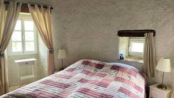 Ein Schlafzimmer mit einem Bett, einem Nachttisch, einer Lampe, einem Fenster mit Vorhängen und einer kleinen Ablage mit einem dekorativen Gegenstand.