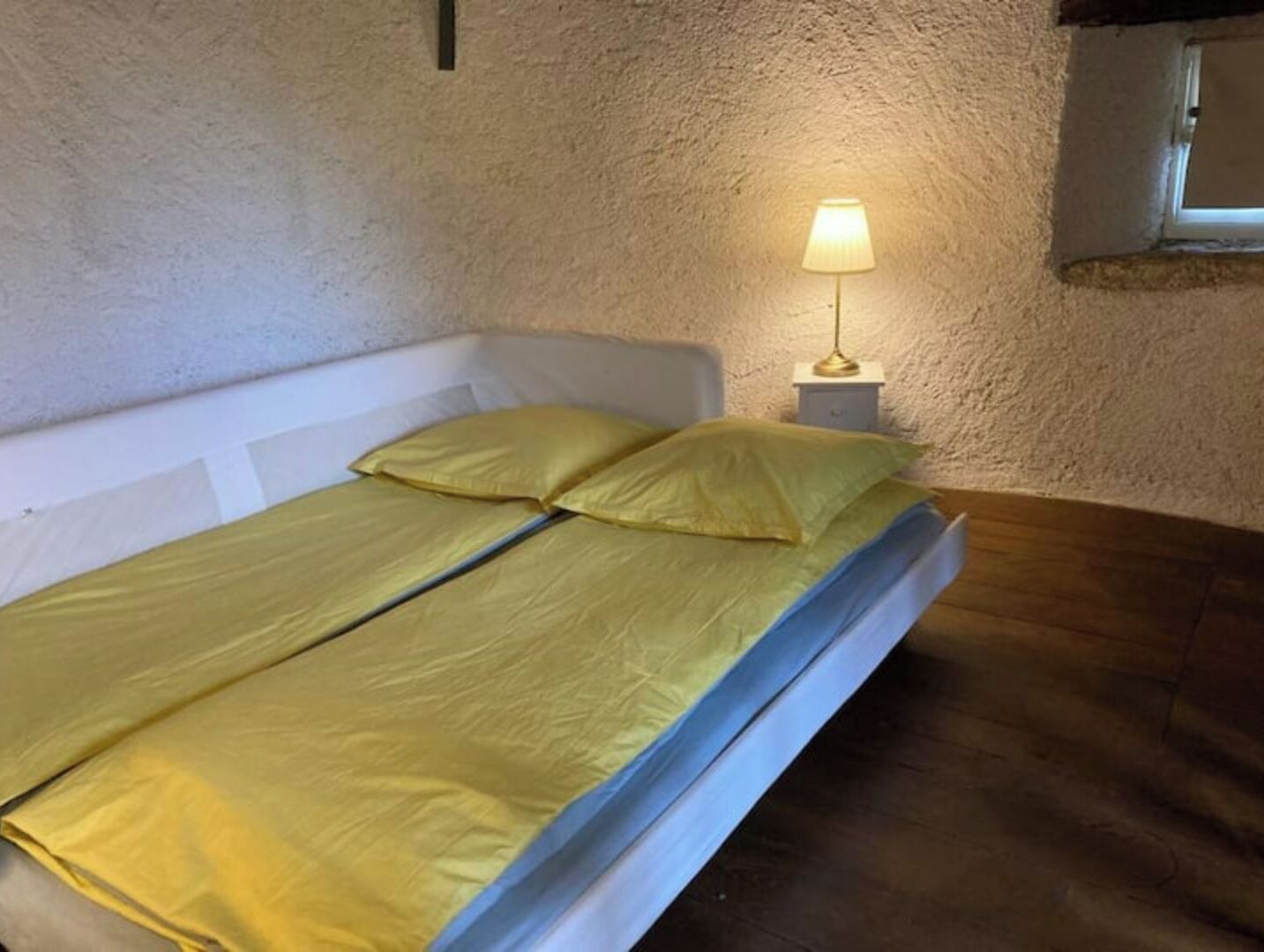 A single bed with a white headboard and a yellow bedspread, a bedside lamp, and a textured wall.