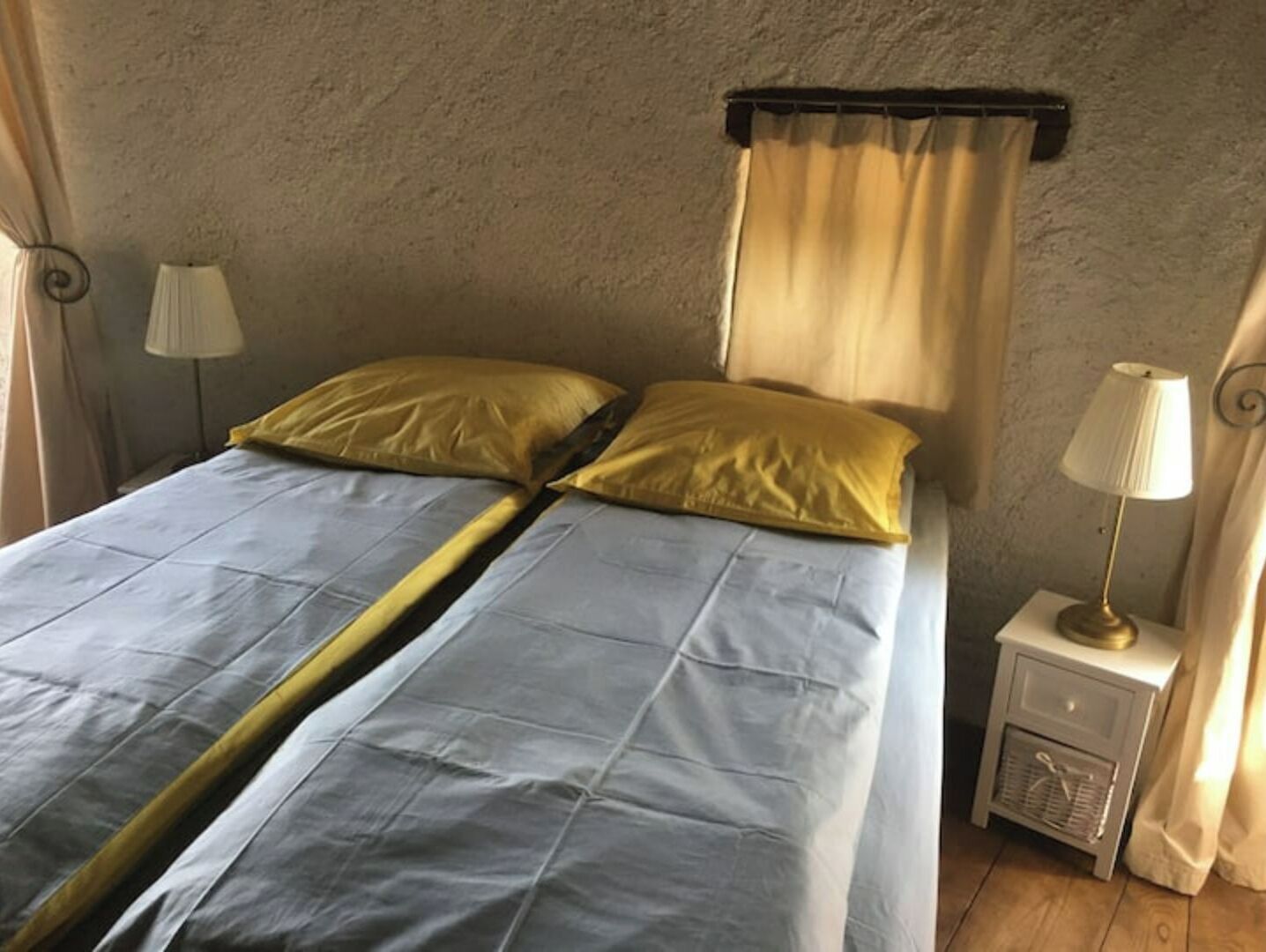 A bed with a blue and yellow bedspread, two pillows, a nightstand with a lamp, and a wall-mounted curtain.