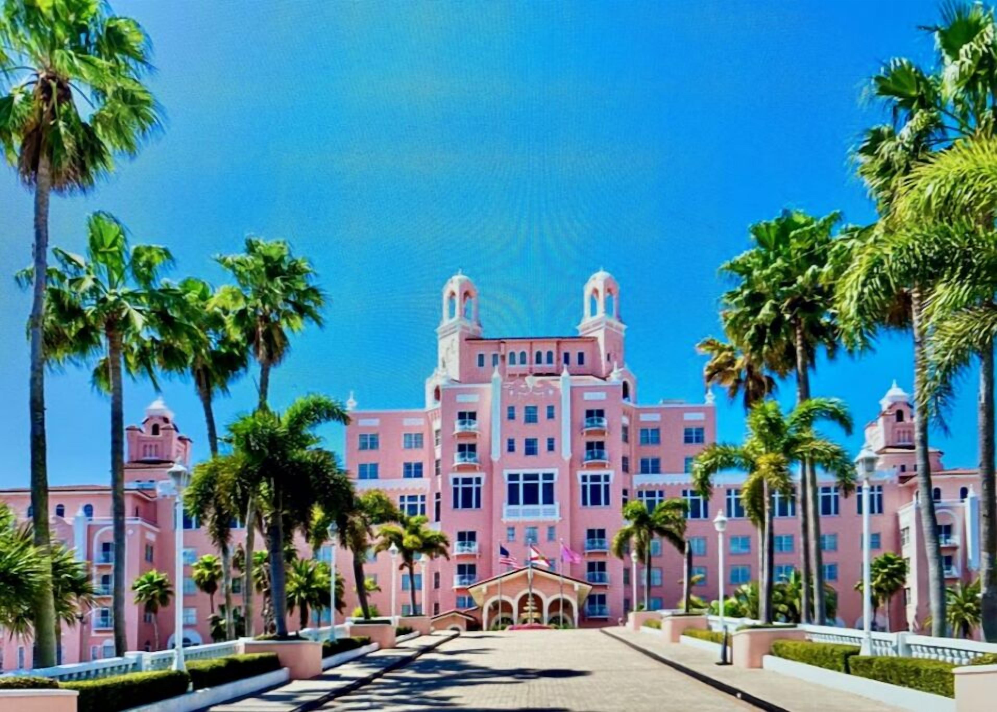Et pink hotel med en central indgang, omgivet af palmer og en klar blå himmel.