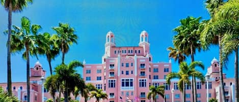 Et pink hotel med en central indgang, omgivet af palmer og en klar blå himmel.