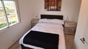 A bedroom with a bed, a nightstand, a bedside table, a painting on the wall, and a window with a view. - Gorgeous 3 Bed En-Suite, Huge Roof Terrace overlooking Pool and La Marquesa Golf (Ciudad Quesada)