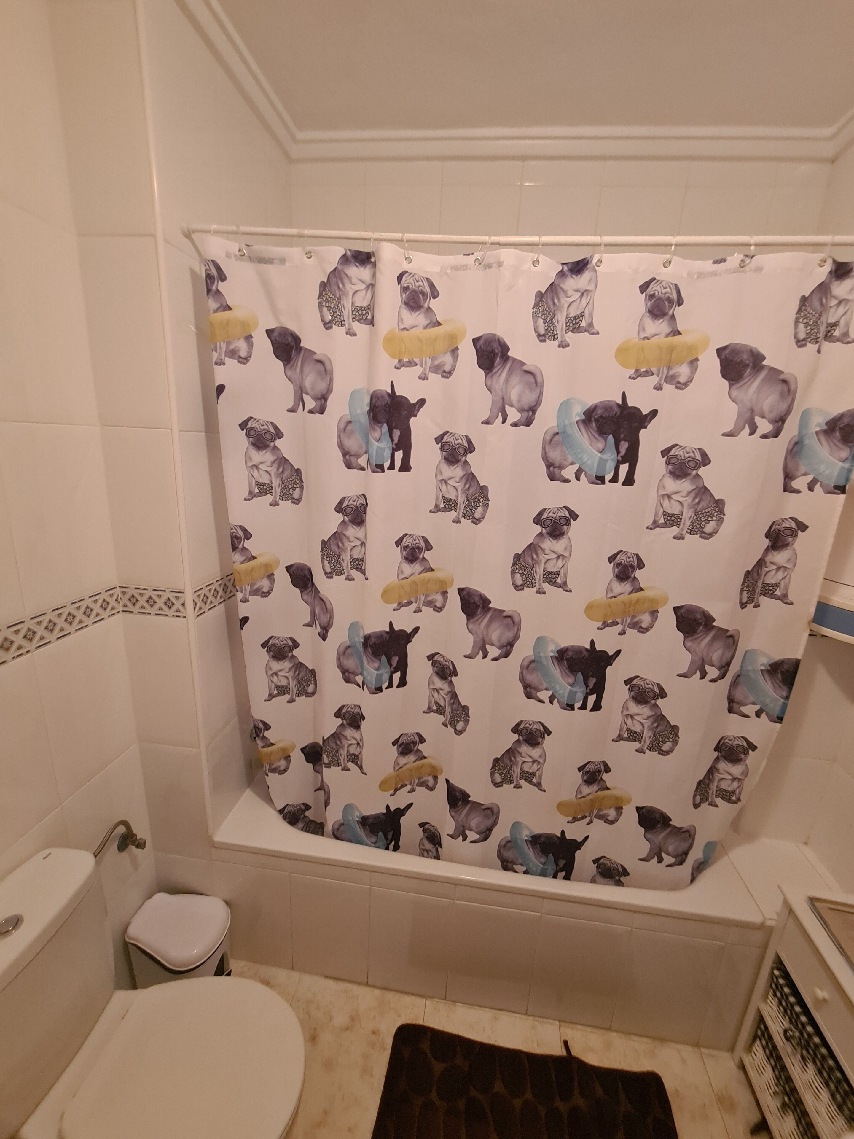 Kylpyhuone, jossa on suihkuverho, jonka kuvio on mopsikoiran kuva, wc ja pieni roskakori.