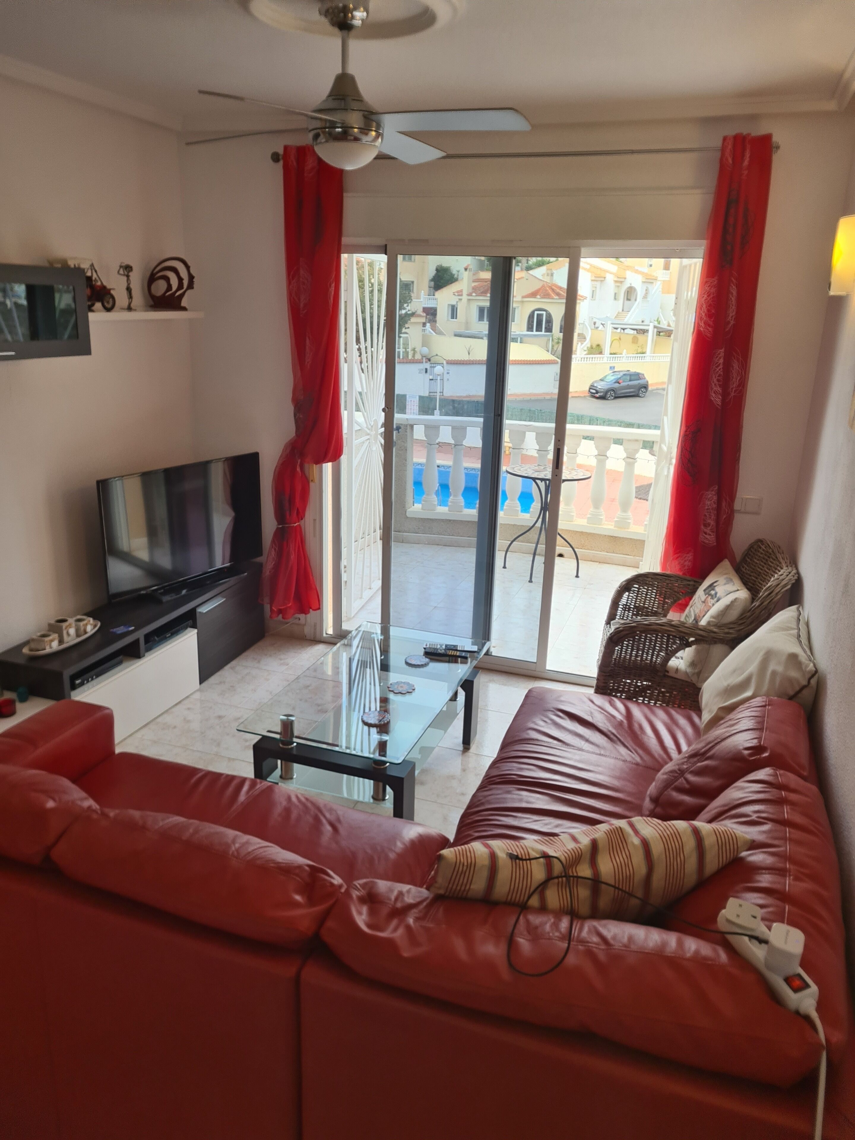 Een woonkamer met een rode bank, een televisie, een glazen salontafel en een balkon met uitzicht op gebouwen en een auto.