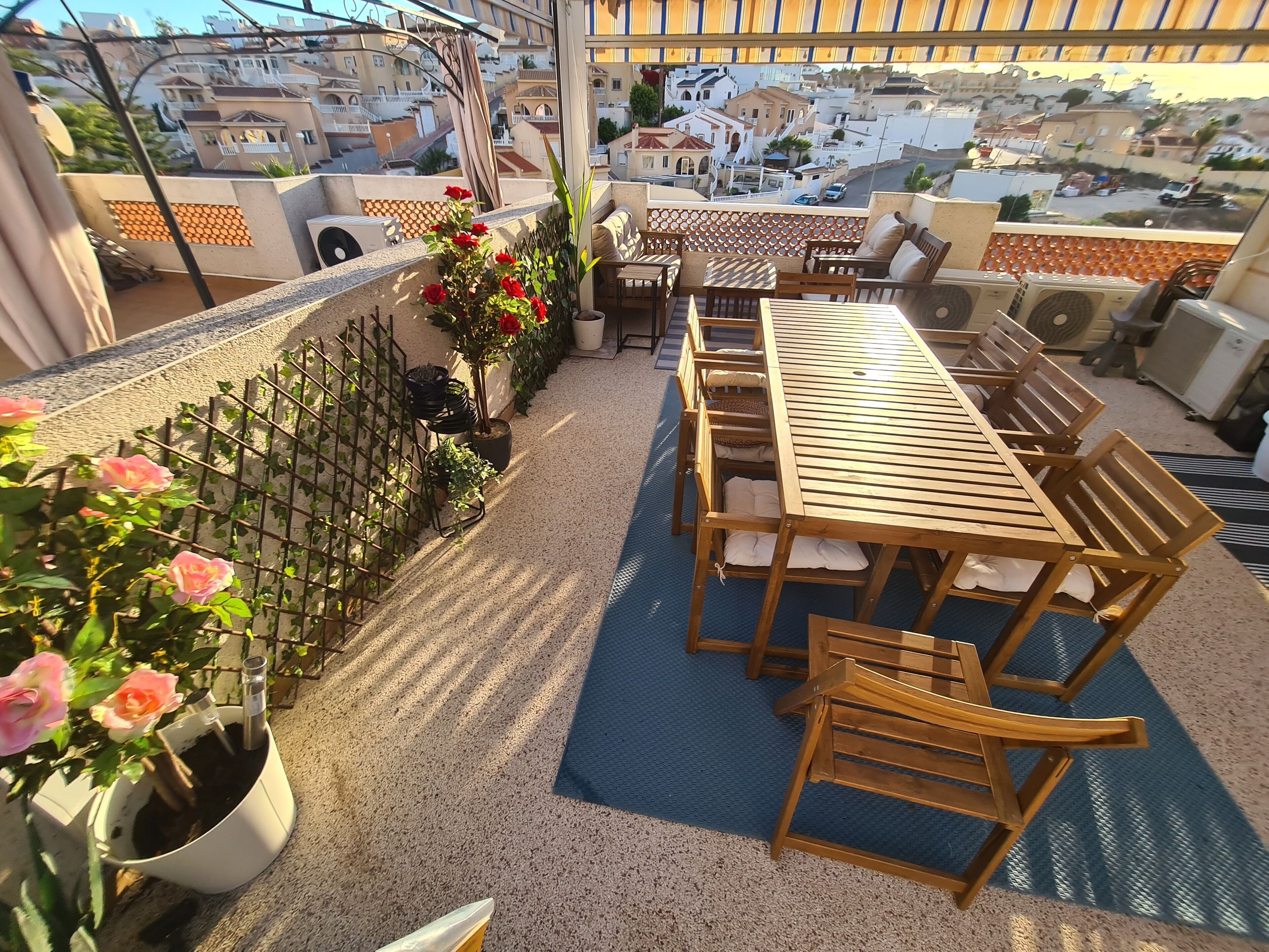 A rooftop terrace with a wooden table and chairs, potted plants, and a view of the cityscape.