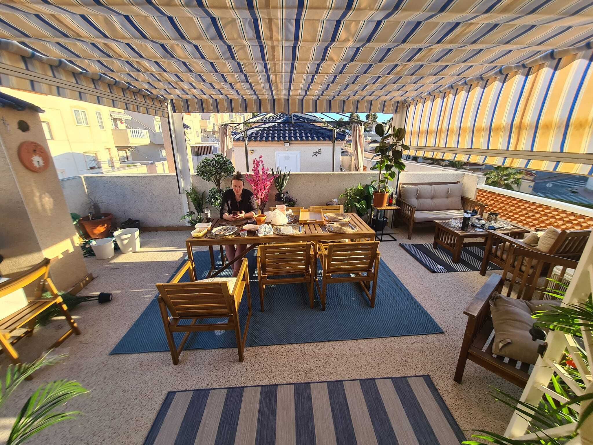 An outdoor patio with a wooden table and chairs, surrounded by potted plants and a striped awning.