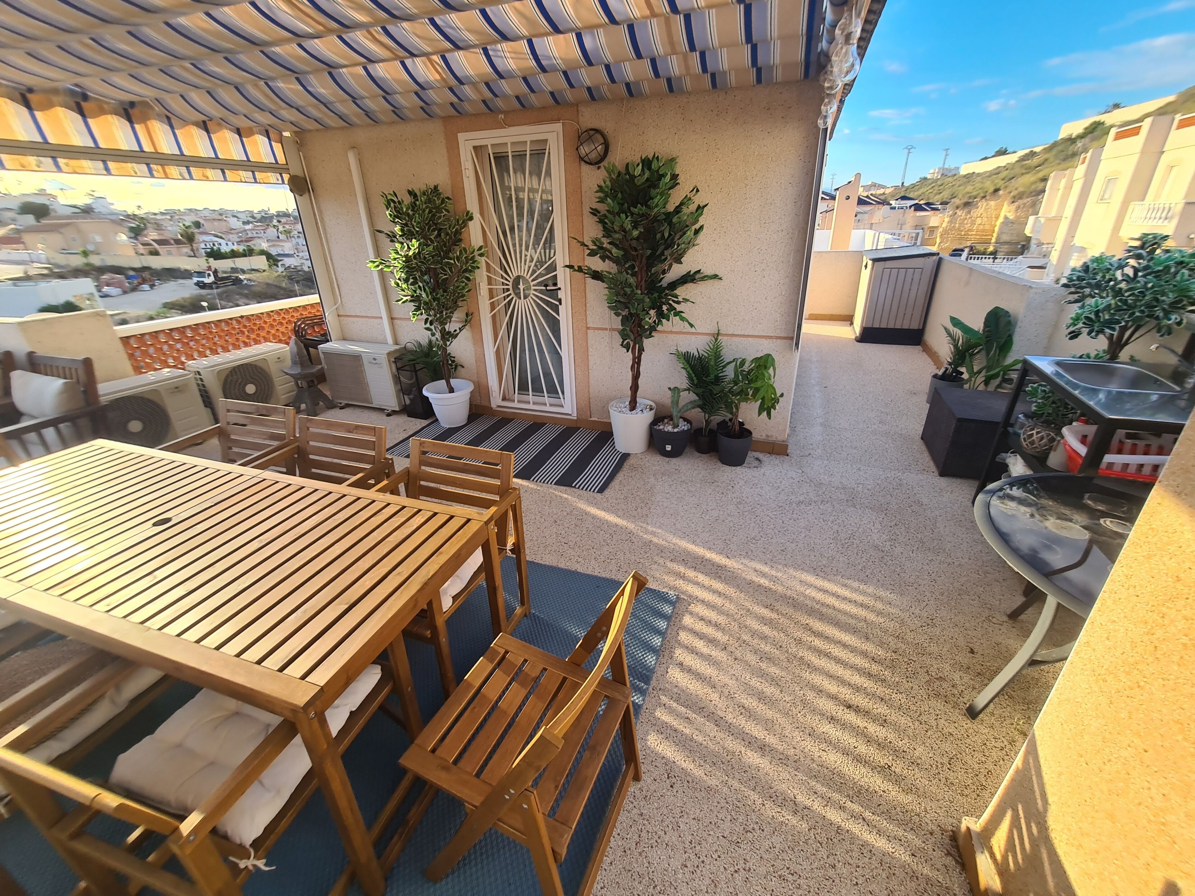 A covered outdoor patio with a wooden table and chairs, potted plants, and a water feature.